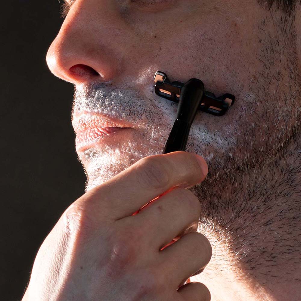 Man holding leaf single edge razor against his face, with shave soap on his beard, closeup.
