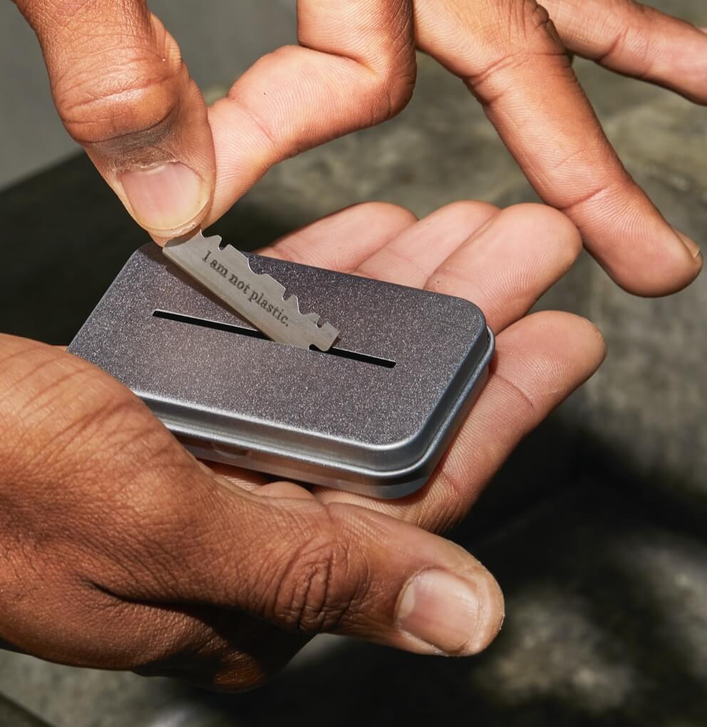 Metal tin with slot on the top, a hand is placing a single edge blade being placed through the slot.