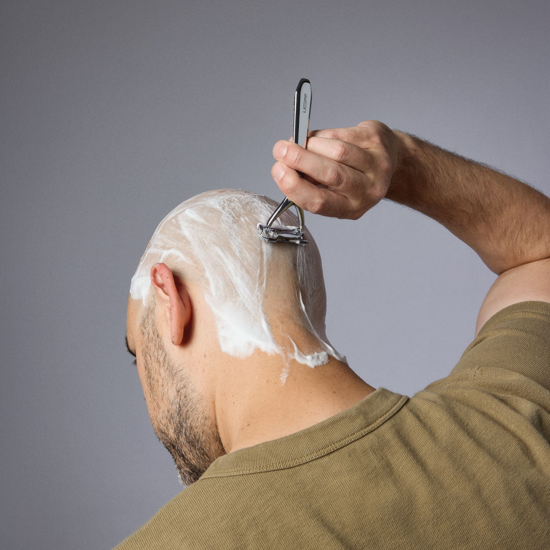 HEAD Person shaving their head with a razor and shaving cream on a gray background.